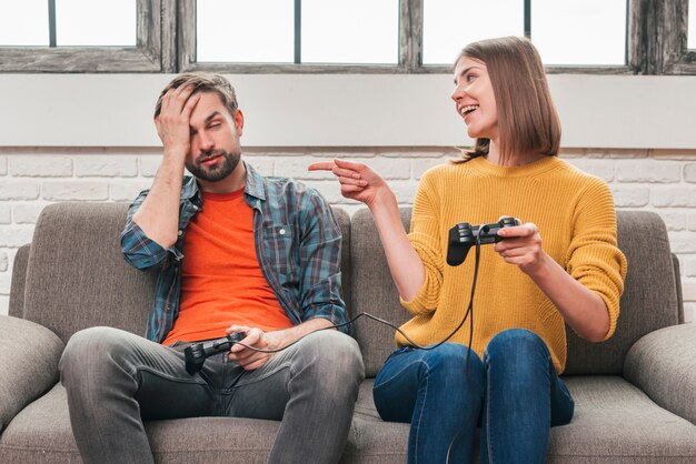 Hombre joven sonriente que se sienta en el sofá que bromea con su novio que sostiene la palanca de mando después de ganar el videojuego