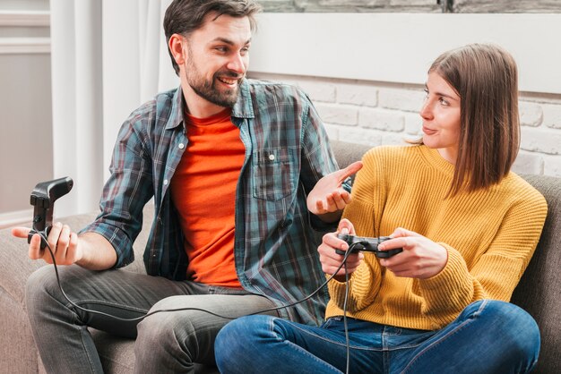 Hombre joven sonriente que se sienta en el sofá que juega con la palanca de mando