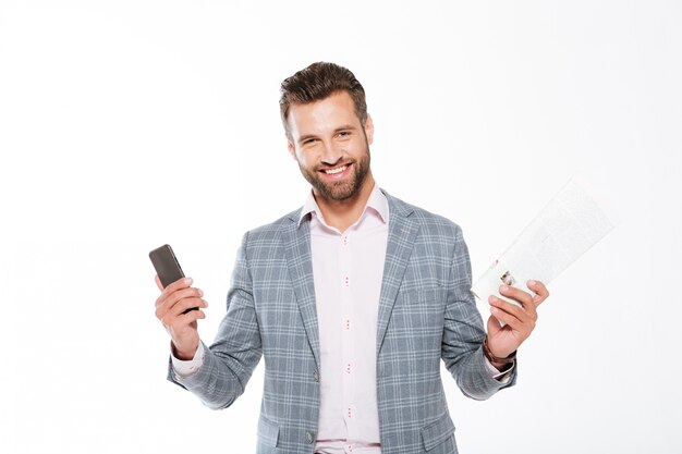 Hombre joven sonriente que sostiene la gaceta y el teléfono móvil.