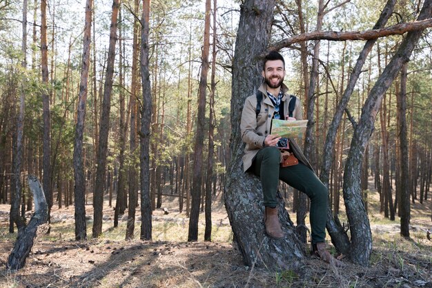 Hombre joven sonriente que sostiene el mapa en la mano que se sienta debajo del árbol en el bosque