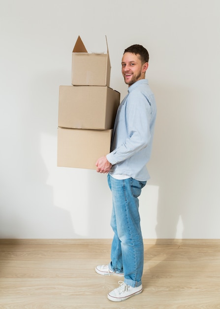 Hombre joven sonriente que sostiene la pila de cajas de cartón en las manos en su nuevo hogar