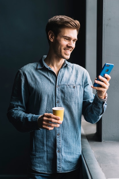 Foto gratuita hombre joven sonriente que sostiene la taza de café para llevar que mira el teléfono móvil