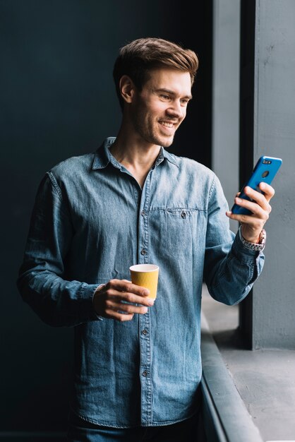 Hombre joven sonriente que sostiene la taza de café para llevar que mira el teléfono móvil