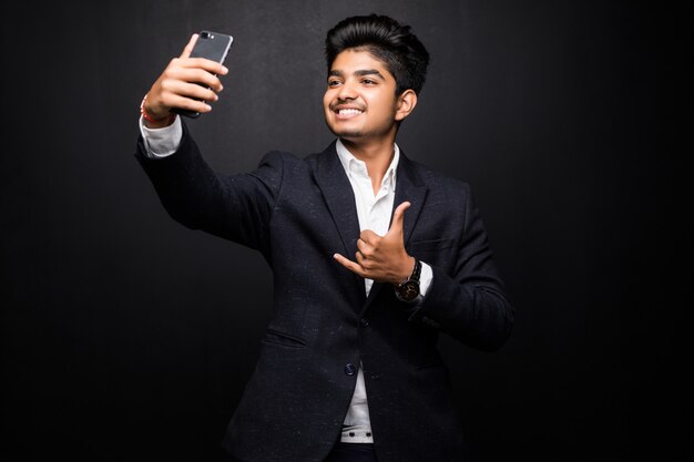 Hombre joven sonriente que toma la foto del selfie en smartphone. Chico indio con dispositivo digital. Concepto de foto selfie. Vista frontal aislada en la pared negra.