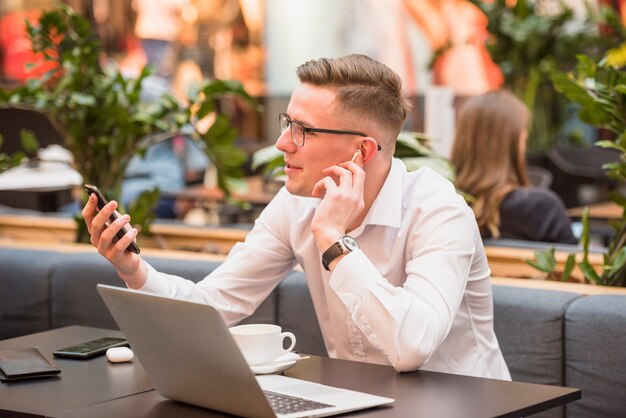 Hombre joven sonriente que usa el auricular inalámbrico que mira el teléfono móvil en caf�