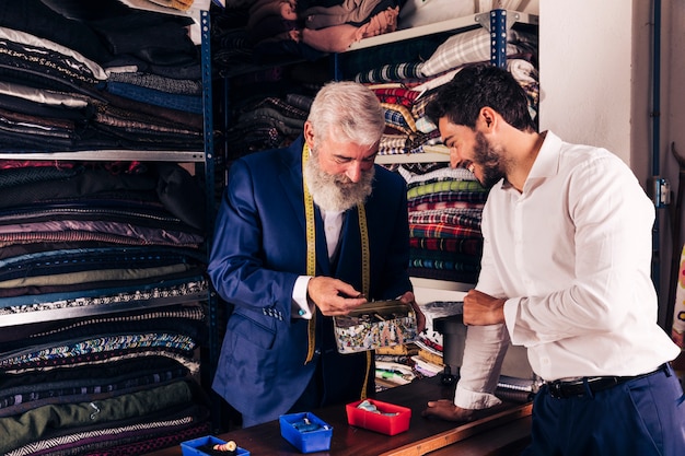 Hombre joven sonriente y sastre mayor que seleccionan el botón del envase en tienda de ropa