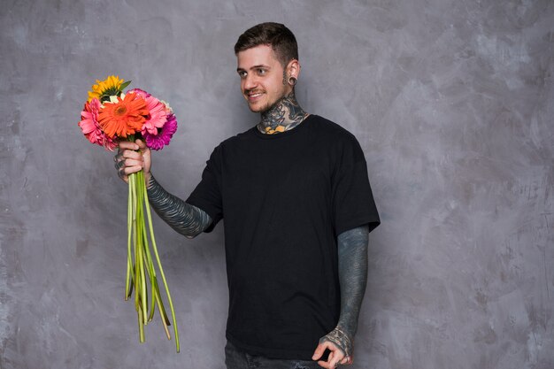 Hombre joven sonriente con el tatuaje en su cuerpo que ofrece las flores del gerbera contra la pared gris