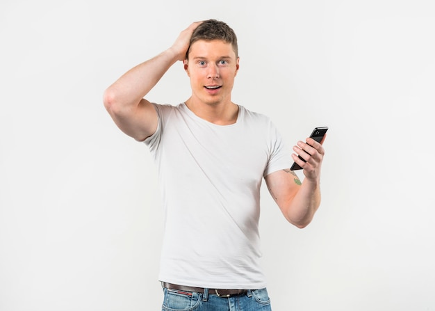 Hombre joven sorprendido que sostiene el teléfono elegante en la mano que mira a la cámara contra el fondo blanco