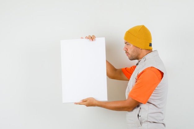 Foto gratuita hombre joven sosteniendo lienzo en blanco en camiseta, chaqueta, sombrero y mirando enfocado.