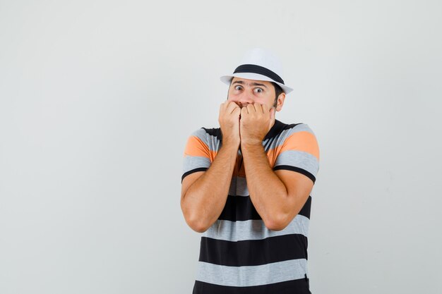 Hombre joven sosteniendo sus puños en la boca con camiseta a rayas, sombrero y mirando asustado. espacio para texto