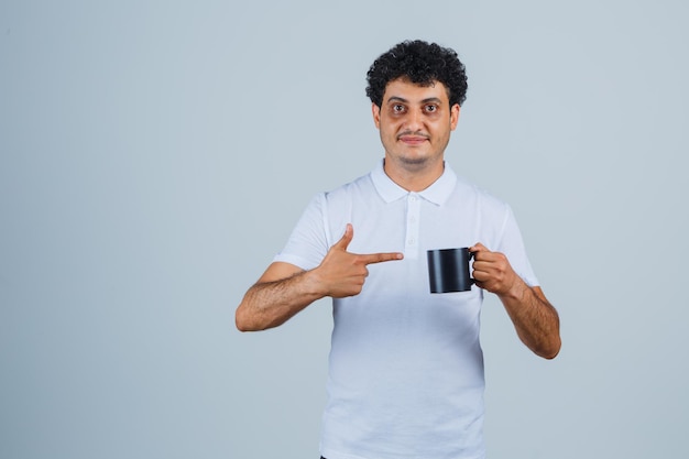 Hombre joven sosteniendo una taza de té mientras apunta hacia ella en camiseta blanca y jeans y parece feliz. vista frontal.