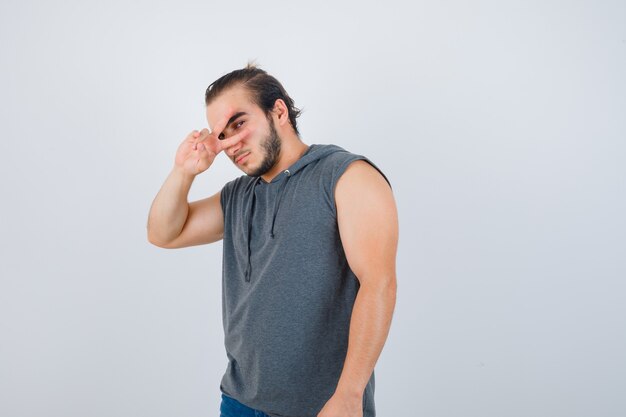 Hombre joven en sudadera con capucha sin mangas que muestra el signo V en el ojo y mirando serio, vista frontal.