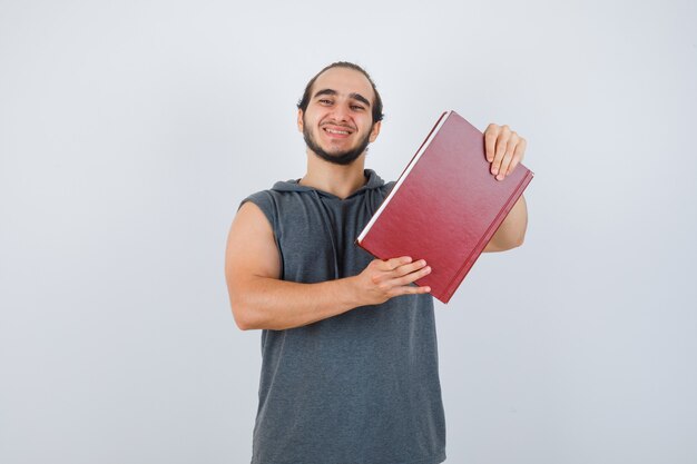 Hombre joven en sudadera con capucha sin mangas que sostiene el libro mientras posa y parece feliz, vista frontal.