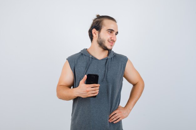Hombre joven en sudadera con capucha sin mangas que sostiene el teléfono móvil mientras mantiene la mano en la cadera y se ve guapo, vista frontal.