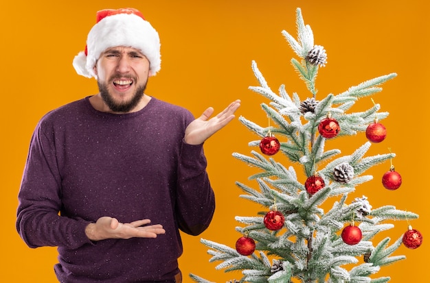 Hombre joven en suéter morado y gorro de Papá Noel de pie junto al árbol de navidad presentándolo con los brazos de las manos mirando confundido y disgustado sobre fondo naranja