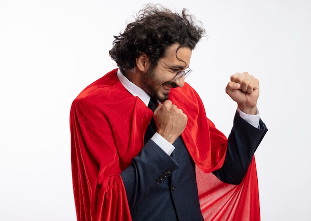 Foto gratuita hombre joven superhéroe alegre en gafas ópticas con traje con capa roja se encuentra de lado manteniendo los puños aislados en la pared blanca