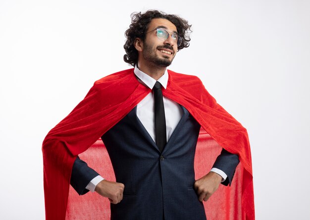 Hombre joven superhéroe complacido con gafas ópticas con traje con capa roja pone las manos en la cintura y mira hacia arriba aislado en la pared blanca
