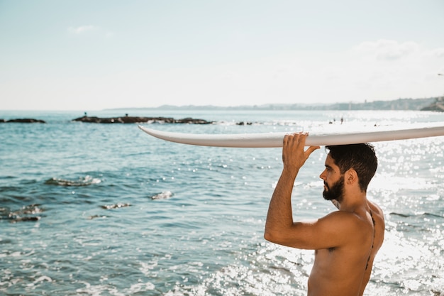 Hombre joven con tabla de surf en la cabeza en la costa cerca del agua