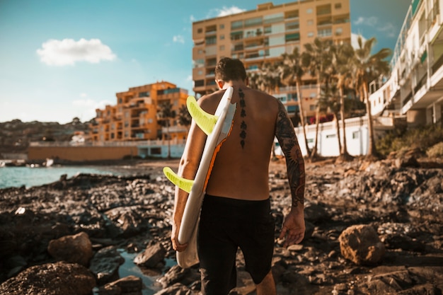 Hombre joven con tabla de surf en la orilla de la roca cerca del agua y edificios