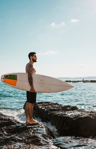 Hombre joven con tabla de surf en roca cerca del agua