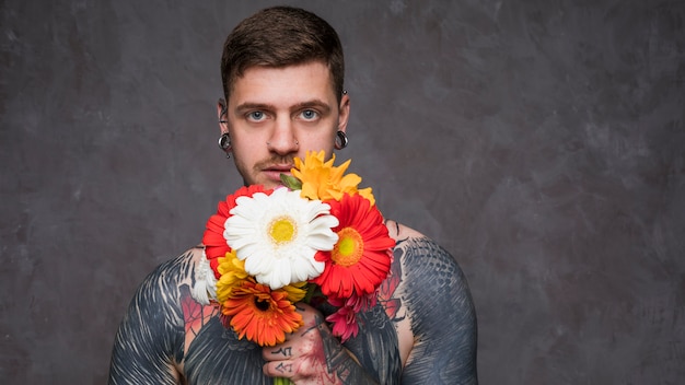 Hombre joven tatuado descamisado con las orejas perforadas que sostienen la flor colorida del gerbera en la mano