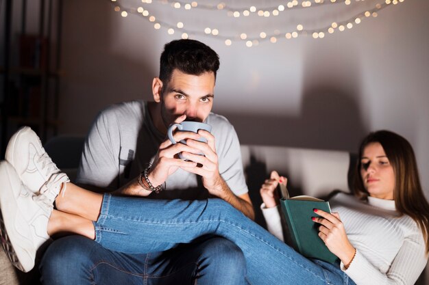 Hombre joven con la taza que ve la TV y la mujer con el libro en el sofá