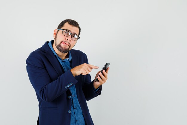 Hombre joven con teléfono móvil, sacando la lengua en camisa, chaqueta y mirando divertido