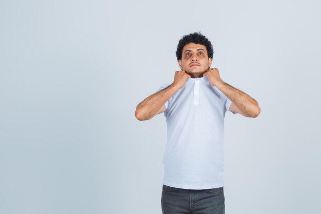 Hombre joven tirando del cuello de su camiseta en camiseta blanca, pantalones y mirando ansioso. vista frontal.