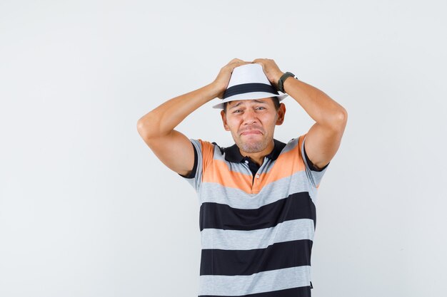 Hombre joven tomados de la mano en la cabeza con camiseta y sombrero y mirando triste