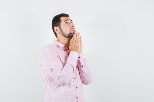 Hombre joven tomados de la mano en gesto de oración en camisa rosa y mirando esperanzado