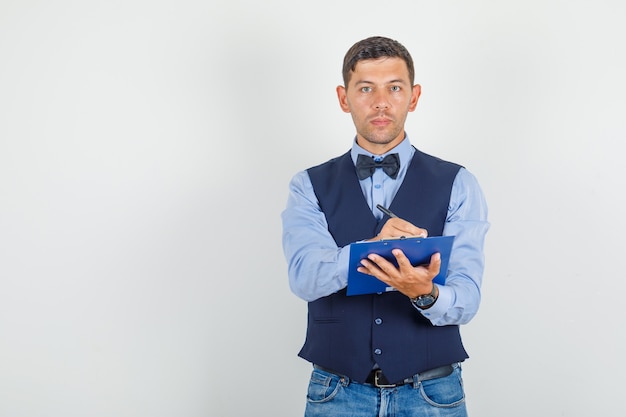 Foto gratuita hombre joven tomando notas en el portapapeles en traje, jeans y mirando serio.