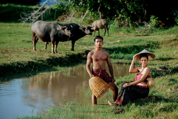 Hombre joven en topless de pie y sosteniendo una trampa de pesca de bambú para pescar peces para cocinar con una hermosa mujer sentada cerca del pantano