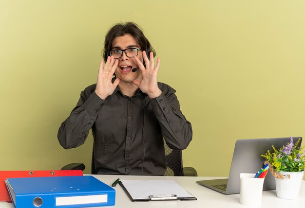 Hombre joven trabajador de oficina alegre en auriculares con gafas ópticas se sienta en el escritorio con herramientas de oficina usando laptop pretende llamar a alguien aislado sobre fondo verde con espacio de copia