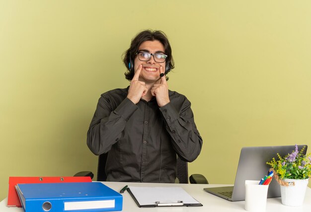 Hombre joven trabajador de oficina en auriculares en gafas ópticas se sienta en el escritorio con herramientas de oficina usando laptop sostiene sonrisa con dedos aislados sobre fondo verde con espacio de copia