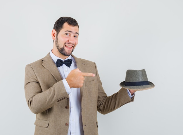 Hombre joven en traje apuntando al sombrero y mirando alegre, vista frontal.