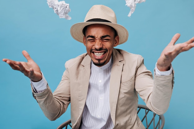 Hombre joven en traje beige esparce sábanas arrugadas sobre fondo azul Chico alegre en chaqueta y camisa blanca posando sobre fondo aislado