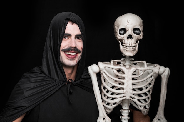 Hombre joven en traje de Halloween posando en estudio con esqueleto