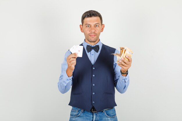 Hombre joven en traje, jeans sosteniendo billetes en euros y jugando a las cartas y mirando contento
