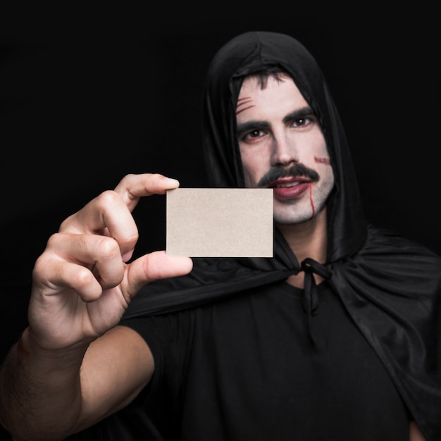 Hombre joven en traje negro de Halloween que muestra poca tarjeta de papel en blanco