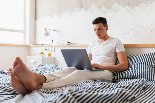 Hombre joven en traje de pijama casual sentado en la cama en la mañana trabajando en la computadora portátil, autónomo ocupado en casa