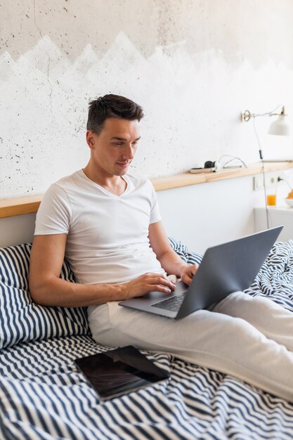 Hombre joven en traje de pijama casual sentado en la cama en la mañana trabajando en la computadora portátil, autónomo ocupado en casa