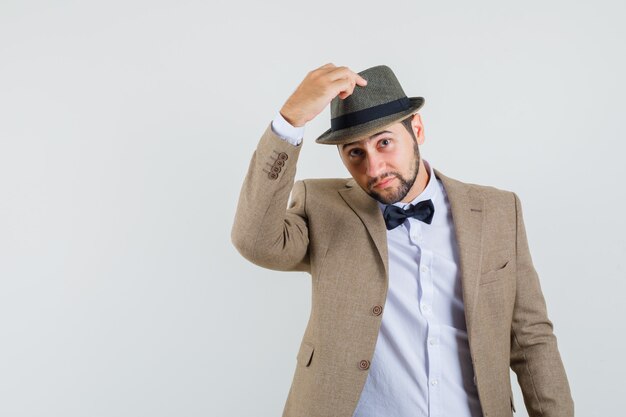 Hombre joven en traje quitándose el sombrero y mirando suave, vista frontal.