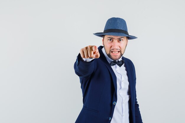 Hombre joven en traje, sombrero apuntando a la cámara y mirando furioso, vista frontal.