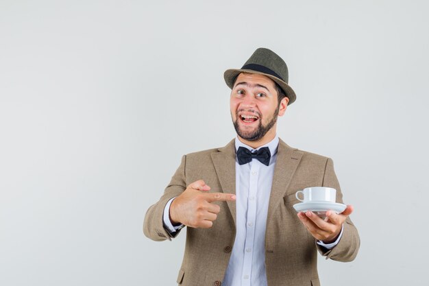 Hombre joven en traje, sombrero apuntando a la taza blanca con platillo y mirando contento, vista frontal.