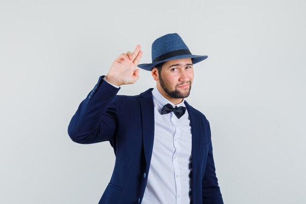 Hombre joven en traje, sombrero gesticulando con dos dedos y mirando genial, vista frontal.