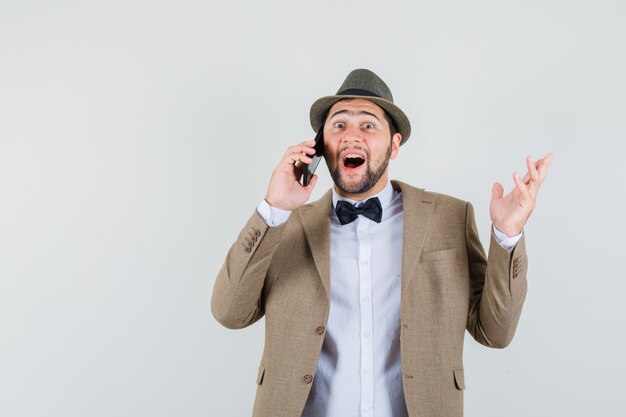 Hombre joven en traje, sombrero hablando por teléfono móvil y mirando feliz, vista frontal.