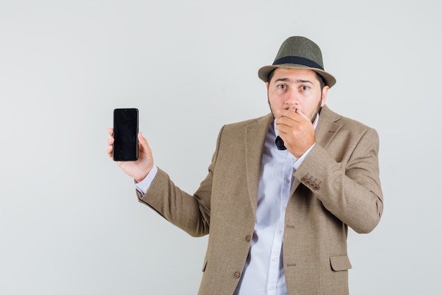 Hombre joven en traje, sombrero mostrando teléfono móvil con la mano en la boca y mirando olvidadizo, vista frontal.