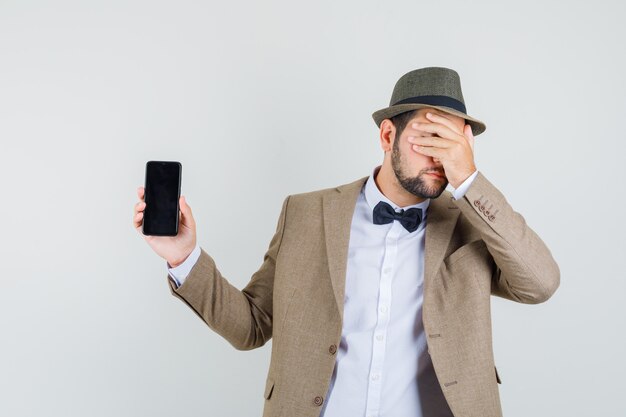 Hombre joven en traje, sombrero mostrando teléfono móvil con la mano en la cara y mirando avergonzado, vista frontal.