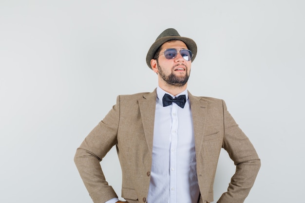 Foto gratuita hombre joven en traje, sombrero de pie con las manos en la cintura y mirando orgulloso, vista frontal.