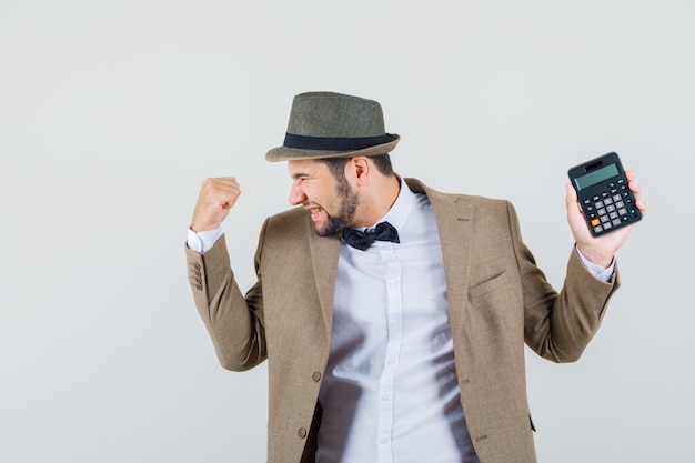 Hombre joven en traje, sombrero sosteniendo calculadora con gesto de ganador y mirando feliz, vista frontal.